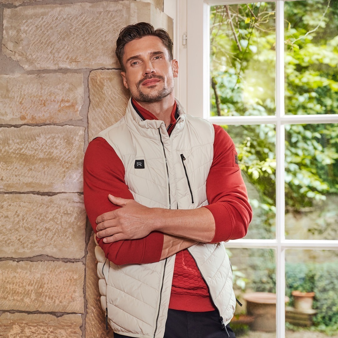 Ein Mann in einem hellen Steppjacke und rotem Pullover steht lässig an einer Wand neben einem Fenster mit Blick auf einen grünen Garten. Ein Mann in einem hellen Steppjacke und rotem Pullover steht lässig an einer Wand neben einem Fenster mit Blick auf einen grünen Garten.