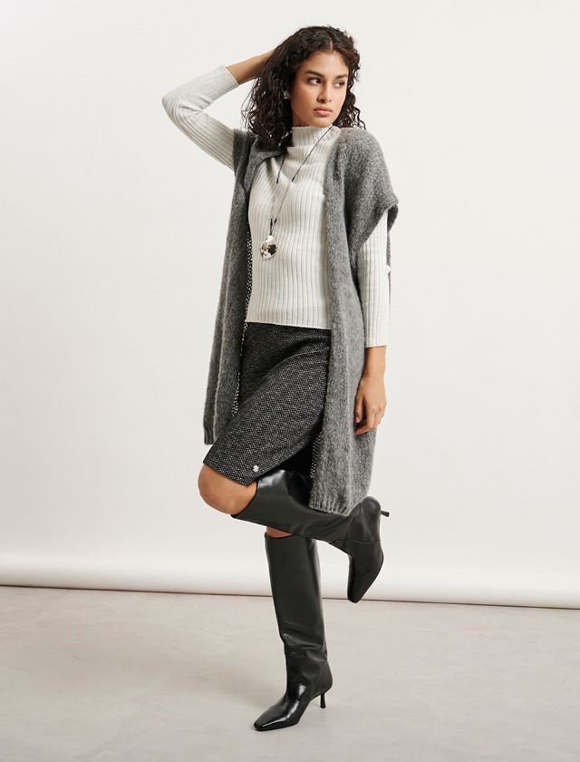 Eine Person präsentiert modische Kleidung: einen grauen Cardigan, ein weißes Oberteil und einen kurzen Rock. Sie trägt hohe, schwarze Stiefel und hat lockige Haare. Eine Person präsentiert modische Kleidung: einen grauen Cardigan, ein weißes Oberteil und einen kurzen Rock. Sie trägt hohe, schwarze Stiefel und hat lockige Haare.