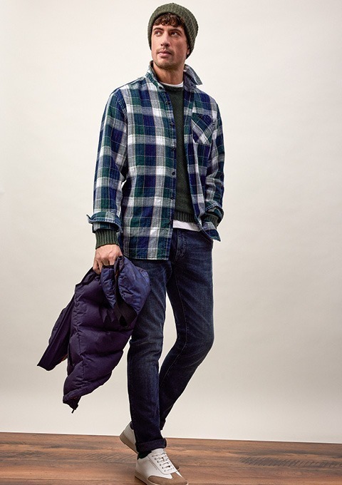 Ein Mann trägt ein kariertes Hemd, eine dunkelgrüne Pullover und eine Mütze, während er eine Jacke in der Hand hält. Ein Mann trägt ein kariertes Hemd, eine dunkelgrüne Pullover und eine Mütze, während er eine Jacke in der Hand hält.