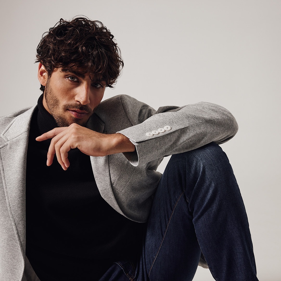 Mann mit lockigem Haar in einem grauen Blazer und schwarzem Pullover, sitzt lässig mit einer Hand an seinem Kinn.r_Marken_76 Mann mit lockigem Haar in einem grauen Blazer und schwarzem Pullover, sitzt lässig mit einer Hand an seinem Kinn.