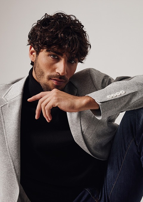 Ein junger Mann mit lockigem Haar trägt einen grauen Blazer und einen schwarzen Pullover. Ein junger Mann mit lockigem Haar trägt einen grauen Blazer und einen schwarzen Pullover.