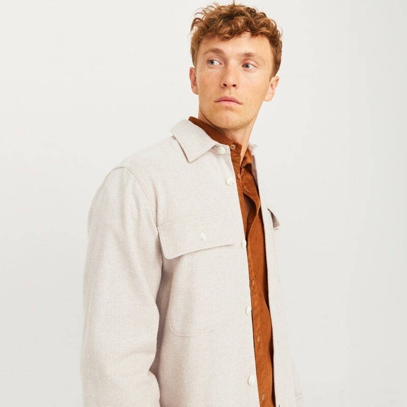 Ein Mann mit lockigem, blonden Haar trägt eine hellbeige Jacke über einem braunen Hemd. Er schaut zur Seite.