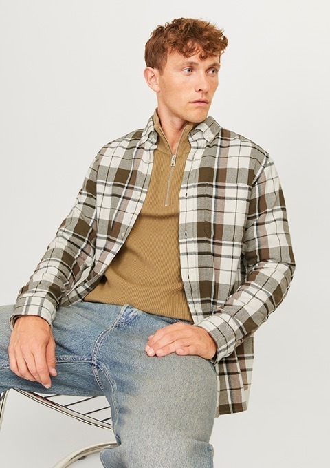 Ein junger Mann sitzt auf einem Stuhl und trägt ein kariertes Hemd sowie einen braunen Pullover.