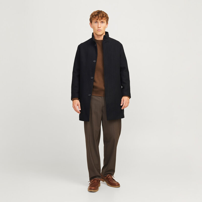 Ein Mann trägt einen schwarzen Mantel, einen braunen Pullover und graue Hosen. Er steht aufrecht vor einem neutralen Hintergrund.