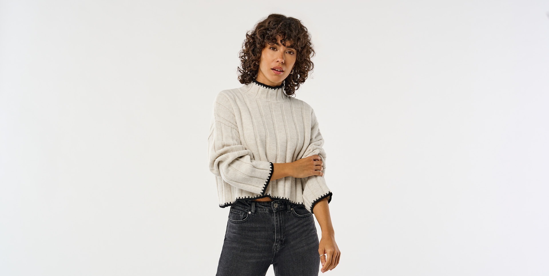 Eine Frau trägt einen hellen, gestreiften Pullover und schwarze Jeans, mit lockigem Haar und einem neutralen Gesichtsausdruck. Eine Frau trägt einen hellen, gestreiften Pullover und schwarze Jeans, mit lockigem Haar und einem neutralen Gesichtsausdruck.