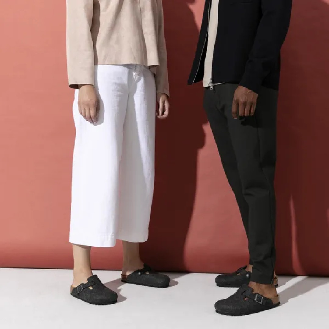 Zwei Personen stehen vor einer roten Wand; die Person links trägt eine helle Bluse und weiße Hose, die Person rechts eine schwarze Jacke und Hose, beide tragen schwarze Schuhe.