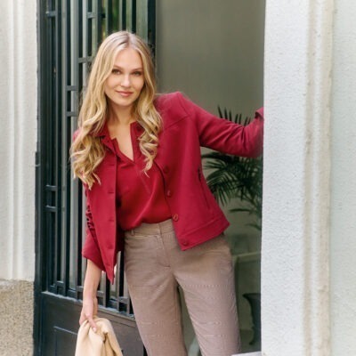 Eine lächelnde Frau mit langen, blonden Locken trägt eine rote Jacke und eine rote Bluse, während sie in einem Eingangsbereich steht.