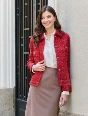 Eine Frau mit langen, braunen Haaren lächelt, gekleidet in eine rote Jacke und einen braunen Rock, steht an einer Wand.