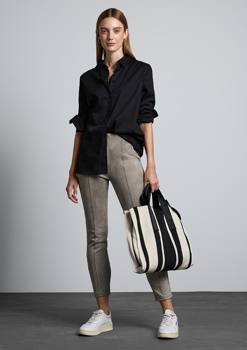 Eine Frau steht in einer modischen Outfit: schwarze Bluse, graue enge Hose und weiße Sneakers. Sie hält eine gestreifte Tasche.