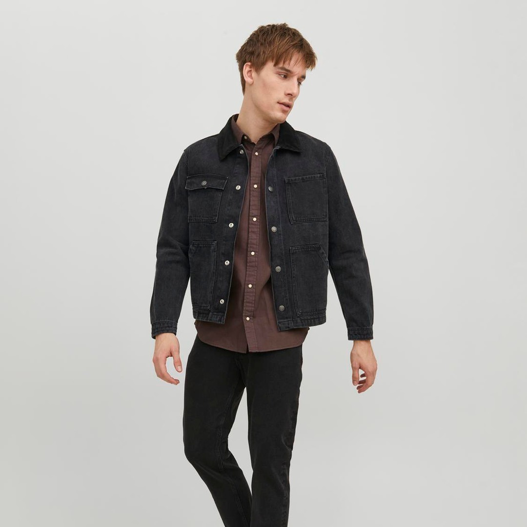 Ein junger Mann trägt eine schwarze Jeansjacke über einem braunen Hemd und steht vor einem neutralen Hintergrund.