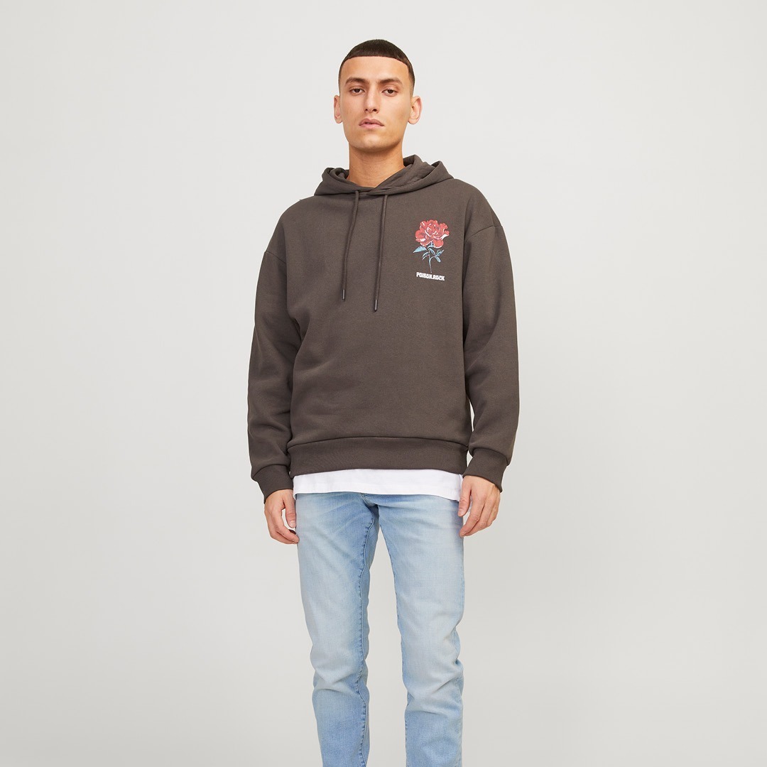Ein junger Mann trägt einen grauen Hoodie mit einer roten Blumen-Illustration und hellblauen Jeans.