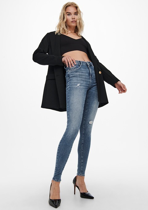 Eine Frau trägt eine schwarze Jacke, ein kurzes Oberteil und enge Jeans. Sie steht in High Heels und posiert.