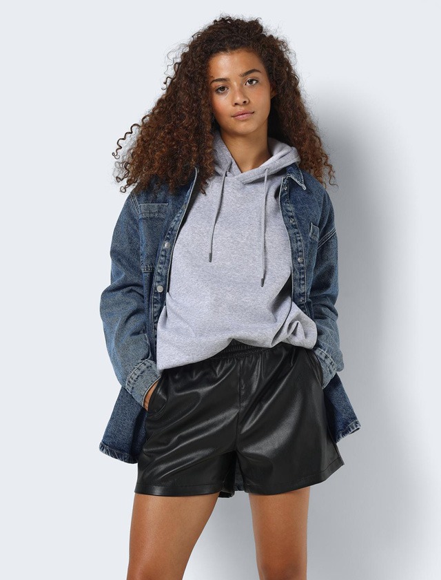 Eine Person mit lockigem Haar trägt ein graues Sweatshirt, eine Jeansjacke und schwarze Leder-Shorts. Eine Person mit lockigem Haar trägt ein graues Sweatshirt, eine Jeansjacke und schwarze Leder-Shorts.