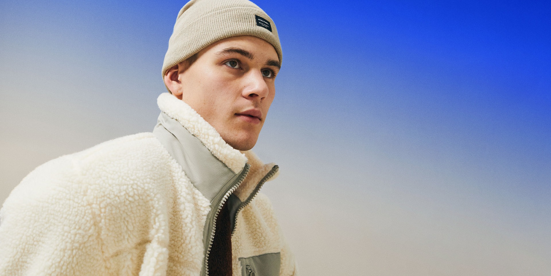 Ein junger Mann trägt eine helle, flauschige Jacke und eine beige Mütze, vor einem blauen Hintergrund. Ein junger Mann trägt eine helle, flauschige Jacke und eine beige Mütze, vor einem blauen Hintergrund.