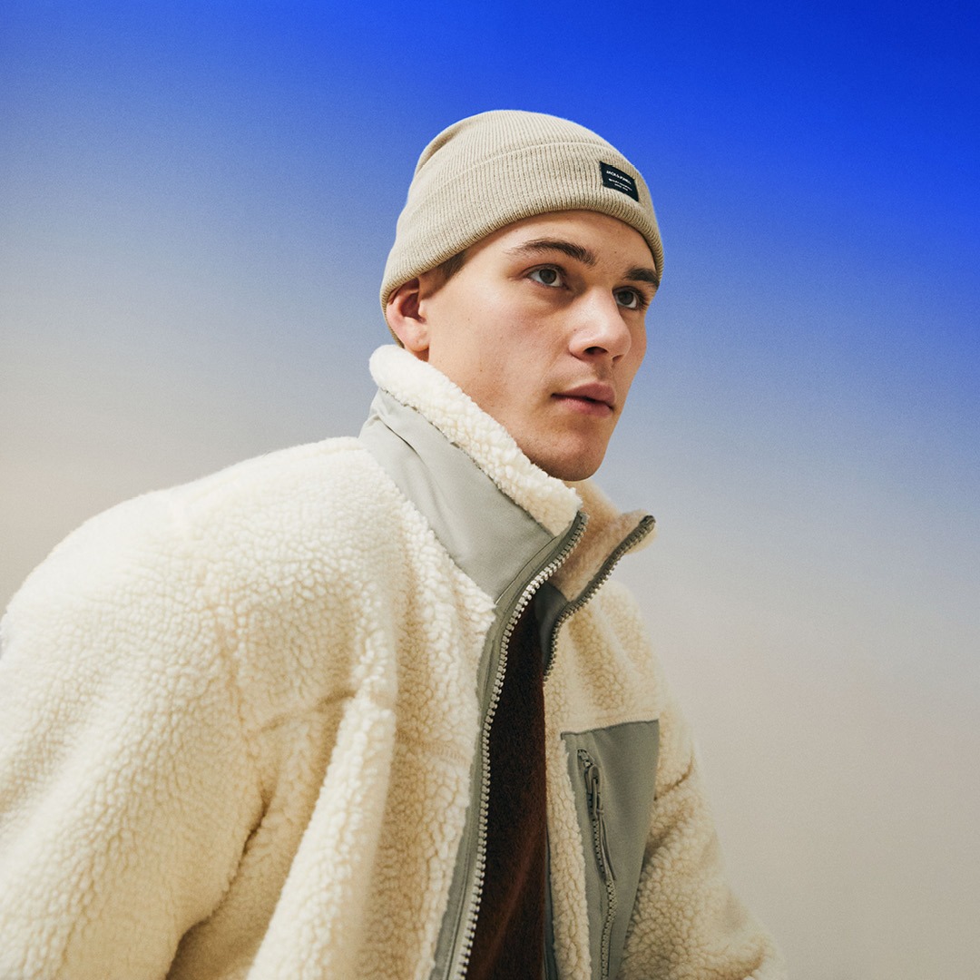 Ein junger Mann trägt eine beige Mütze und eine weiße Fleecejacke mit grauen Akzenten. Der Hintergrund zeigt einen Farbverlauf von Blau zu Beige. Ein junger Mann trägt eine beige Mütze und eine weiße Fleecejacke mit grauen Akzenten. Der Hintergrund zeigt einen Farbverlauf von Blau zu Beige.