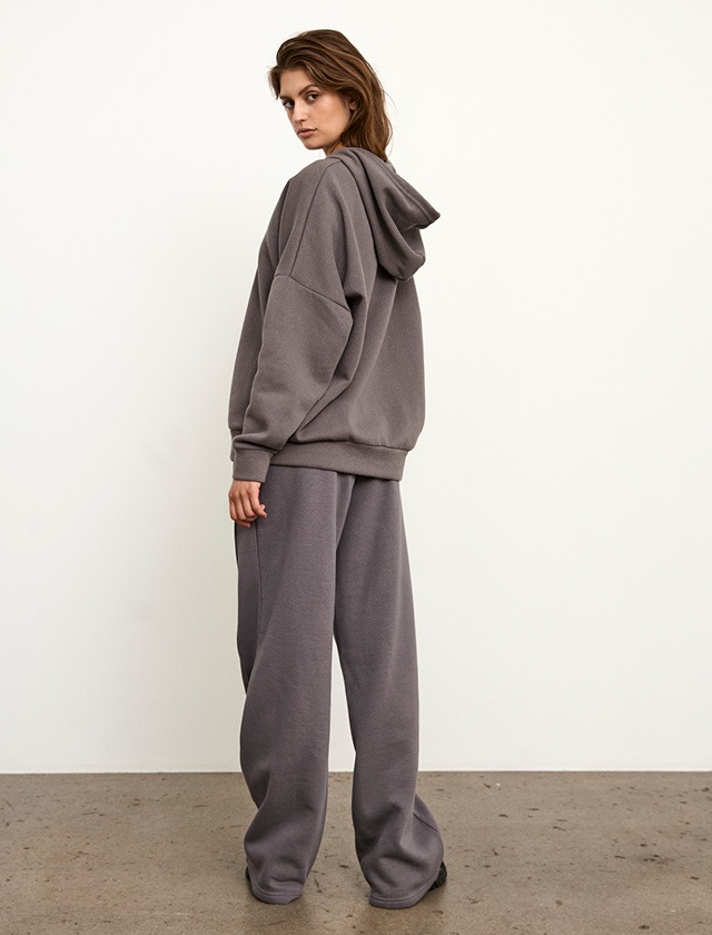 Eine Person trägt einen grauen Hoodie und weite graue Hosen, steht seitlich vor einer weißen Wand. Eine Person trägt einen grauen Hoodie und weite graue Hosen, steht seitlich vor einer weißen Wand.