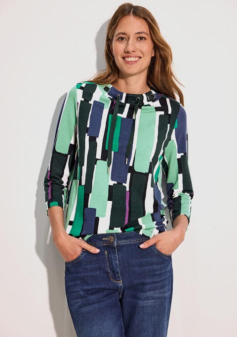 Eine Frau lächelt, trägt ein gestreiftes Oberteil in Grün-, Blau- und Weißtönen und Jeans.