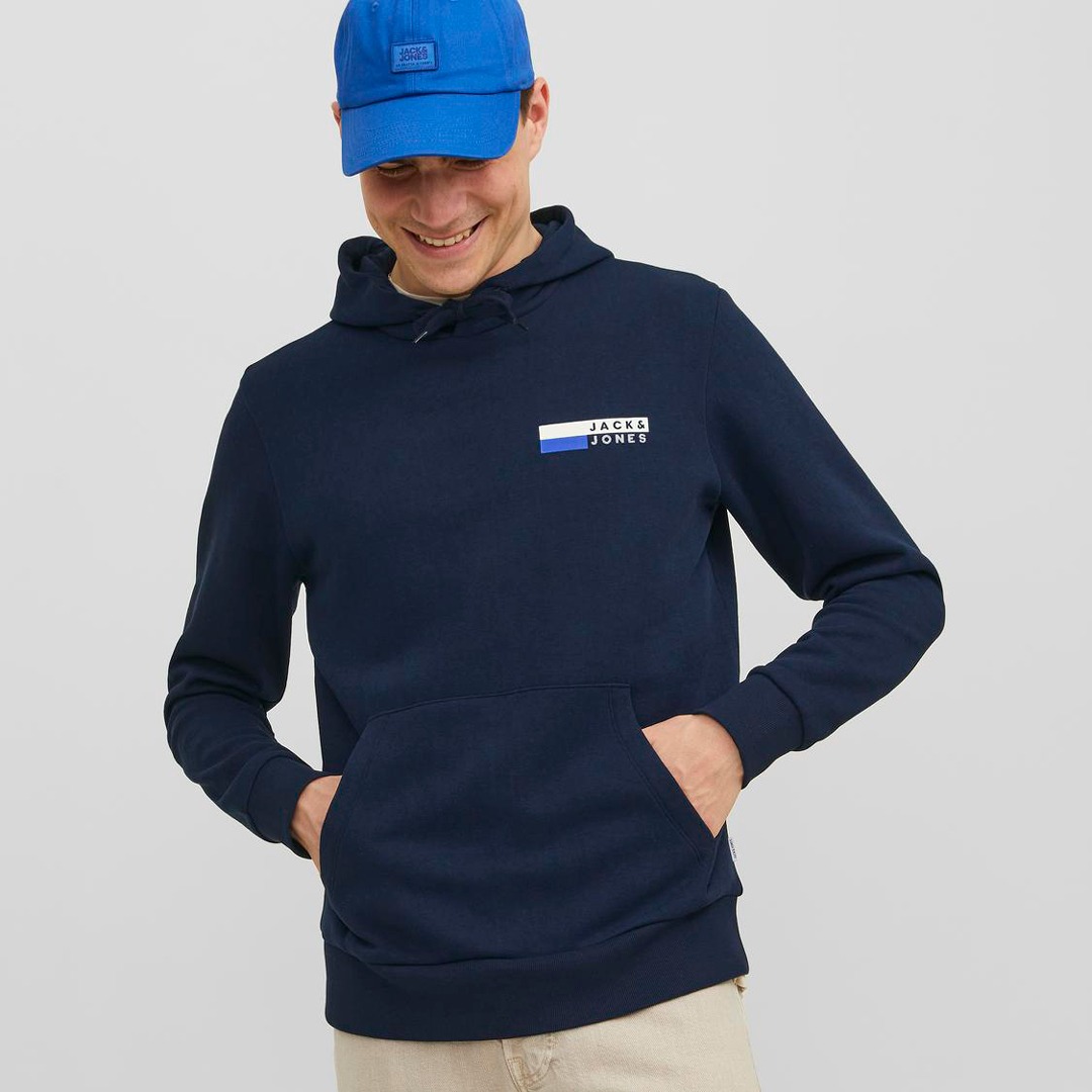 Ein lächelnder Mann trägt einen dunkelblauen Hoodie mit einem Logo und eine blaue Cap.