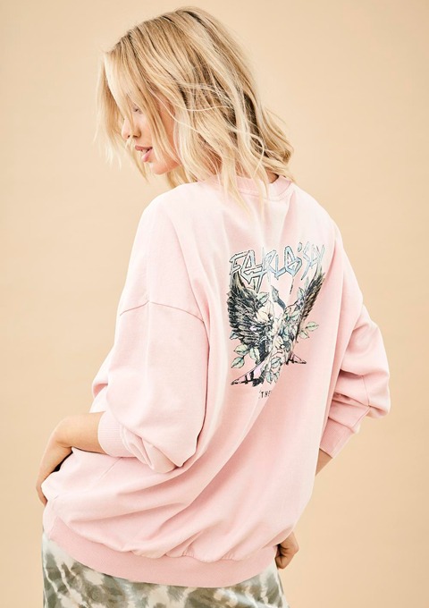 Eine Person trägt einen rosa Pullover mit einem grafischen Druck auf dem Rücken. Der Hintergrund ist beige.