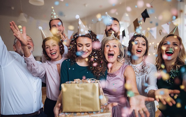 Eine fröhliche Gruppe von Menschen feiert mit Konfetti und Partyhüten, während sie um einen Geschenktisch stehen.