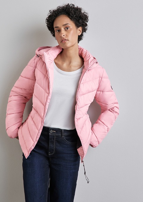 Eine Person trägt eine rosa, gepolsterte Jacke und ein weißes T-Shirt, stehend vor einer grauen Wand. Eine Person trägt eine rosa, gepolsterte Jacke und ein weißes T-Shirt, stehend vor einer grauen Wand.