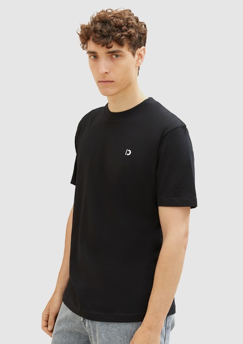 Junger Mann trägt ein schwarzes T-Shirt mit einem kleinen Logo auf der Brust.