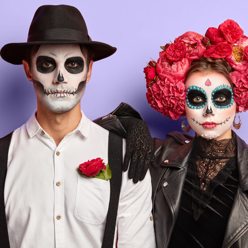 Ein Paar mit Totenschädel-Make-up: ein Mann in weißem Hemd und schwarzem Hut, und eine Frau mit bunten Blumen im Haar und schwarzer Jacke.