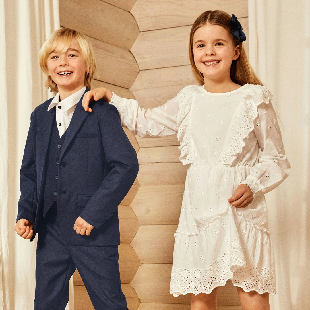 Ein lächelnder Junge in einem dunklen Anzug und ein Mädchen in einem weißen Kleid mit Rüschendetails, die sich in einem hellen Raum mit großen Fenstern befinden.