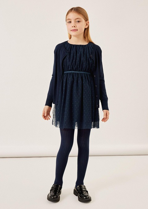 Ein Mädchen trägt ein dunkelblaues Kleid mit einem Oberteil und einem Rock aus transparenter Spitze, dazu eine passende Strickjacke und gestreifte Strumpfhosen.