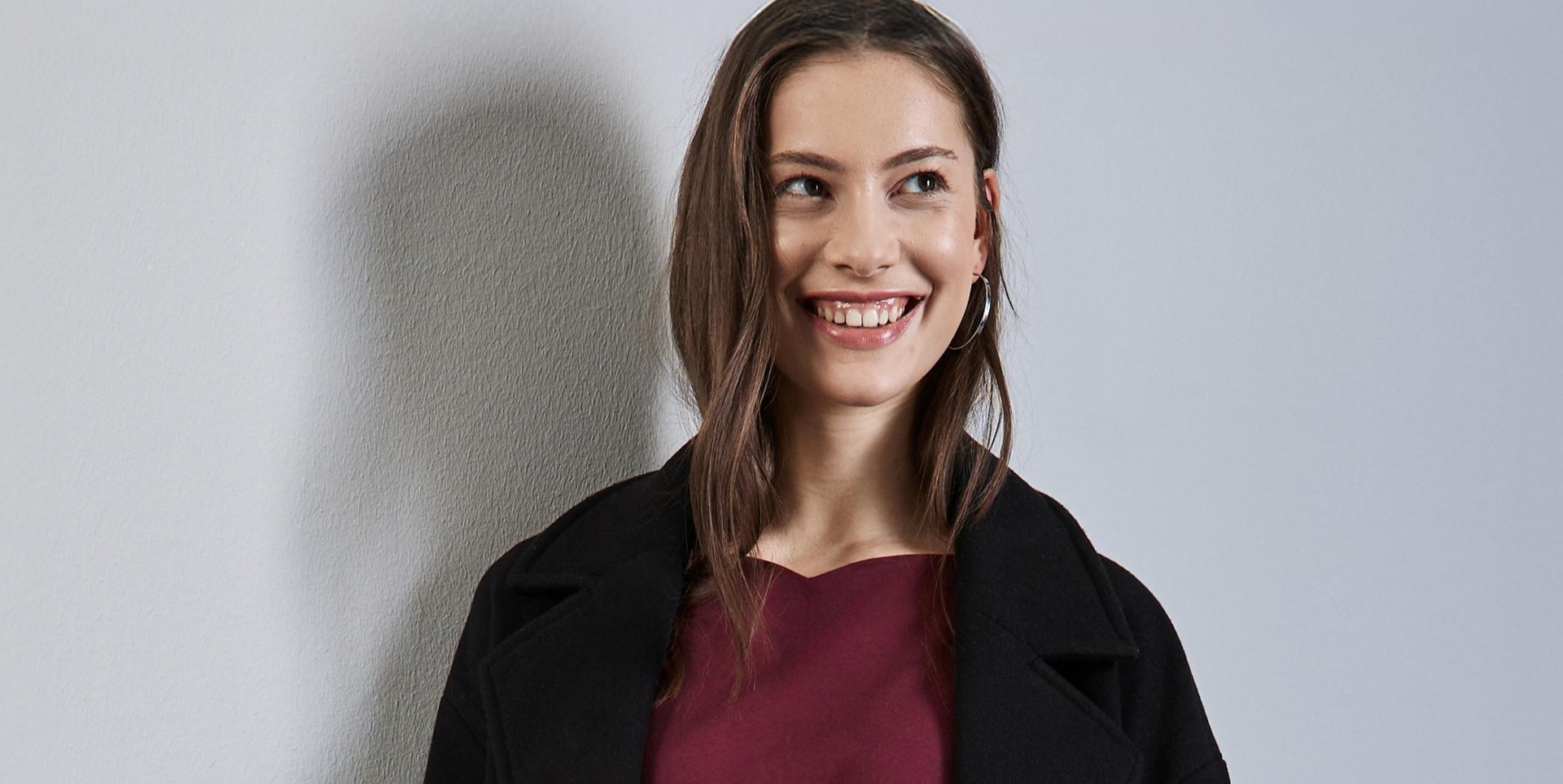 Eine lächelnde Person mit langen Haaren trägt ein dunkles Oberteil und eine schwarze Jacke vor einem grauen Hintergrund. Eine lächelnde Person mit langen Haaren trägt ein dunkles Oberteil und eine schwarze Jacke vor einem grauen Hintergrund.