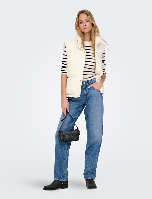 Eine blonde Frau trägt eine gestreifte Bluse, eine weiße Weste, hochwertige Jeans und schwarze Stiefel. Sie hält eine kleine schwarze Tasche.