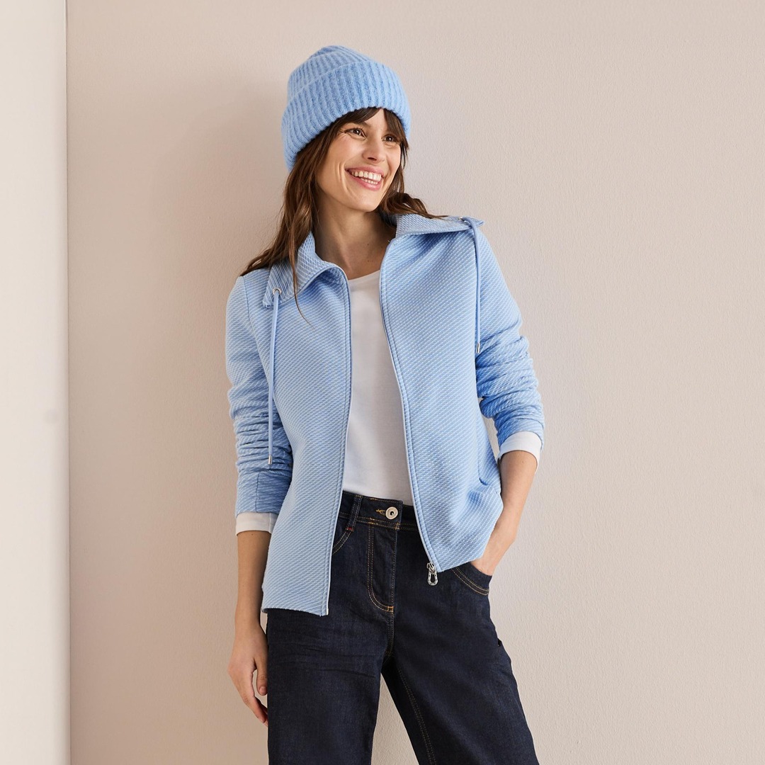 Eine Frau in blauer Jacke und Hut posiert fröhlich, mit einem strahlenden Lächeln.