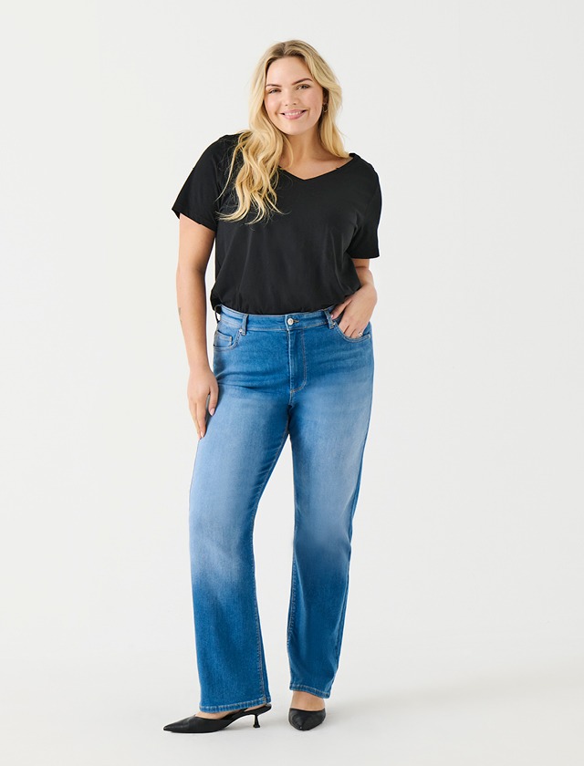 Eine Person trägt ein schwarzes T-Shirt und hellblaue Jeans. Sie lächelt und steht aufrecht.