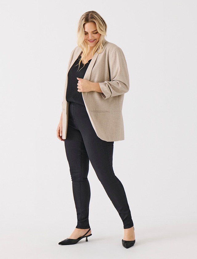 Eine Frau steht in einem beige-farbenen Blazer, schwarzem Oberteil und schwarzen Skinny Jeans. Sie trägt schicke schwarze Schuhe und hat lange, blonde Haare.