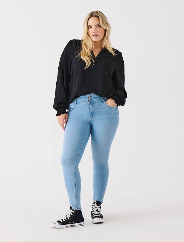 Eine Frau trägt eine schwarze Bluse, hellblaue Skinny-Jeans und schwarze Sneaker. Sie steht auf einem weißen Hintergrund.