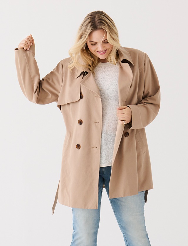 Eine lächelnde Frau trägt einen beigen Trenchcoat über einem hellen Pullover und Jeans.