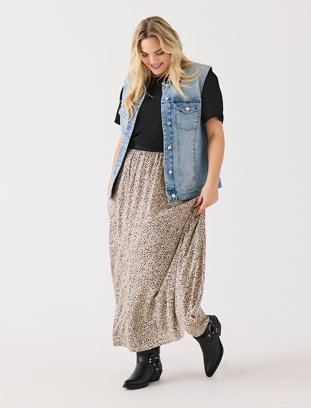 Eine Person trägt eine Jeansweste, ein schwarzes T-Shirt und einen langen Rock mit Leopardenmuster. Sie steht auf einem weißen Hintergrund.