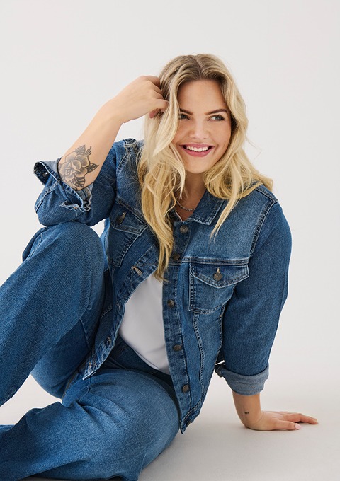 Eine lächelnde Frau mit langen blonden Haaren sitzt in einem blauen Denim-Outfit und berührt ihr Haar.