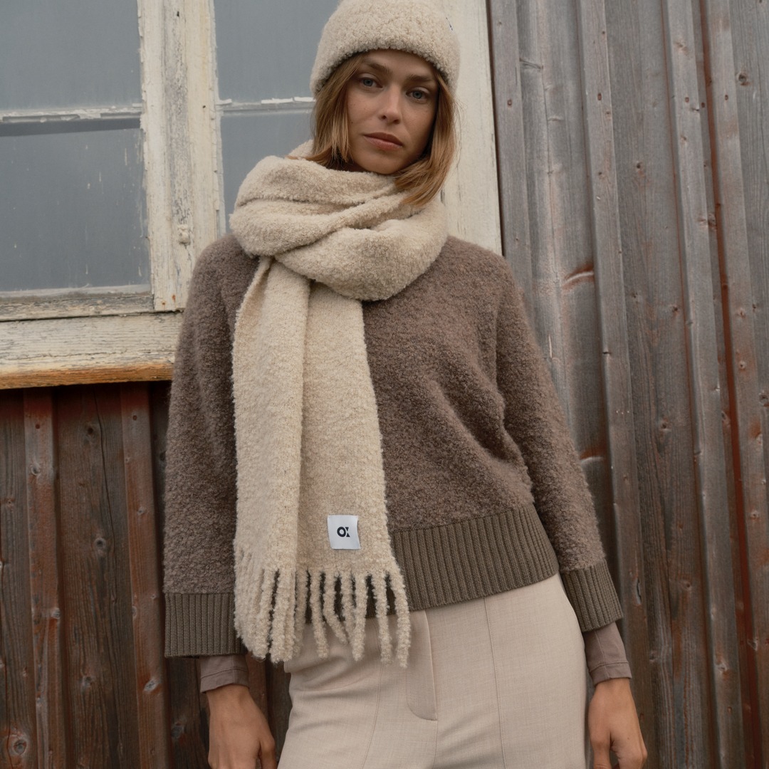 Eine Frau trägt einen braunen pullover, einen beigen Schal und eine bequeme Mütze. Sie steht vor einer Holzfassade und blickt in die Kamera. Eine Frau trägt einen braunen pullover, einen beigen Schal und eine bequeme Mütze. Sie steht vor einer Holzfassade und blickt in die Kamera.