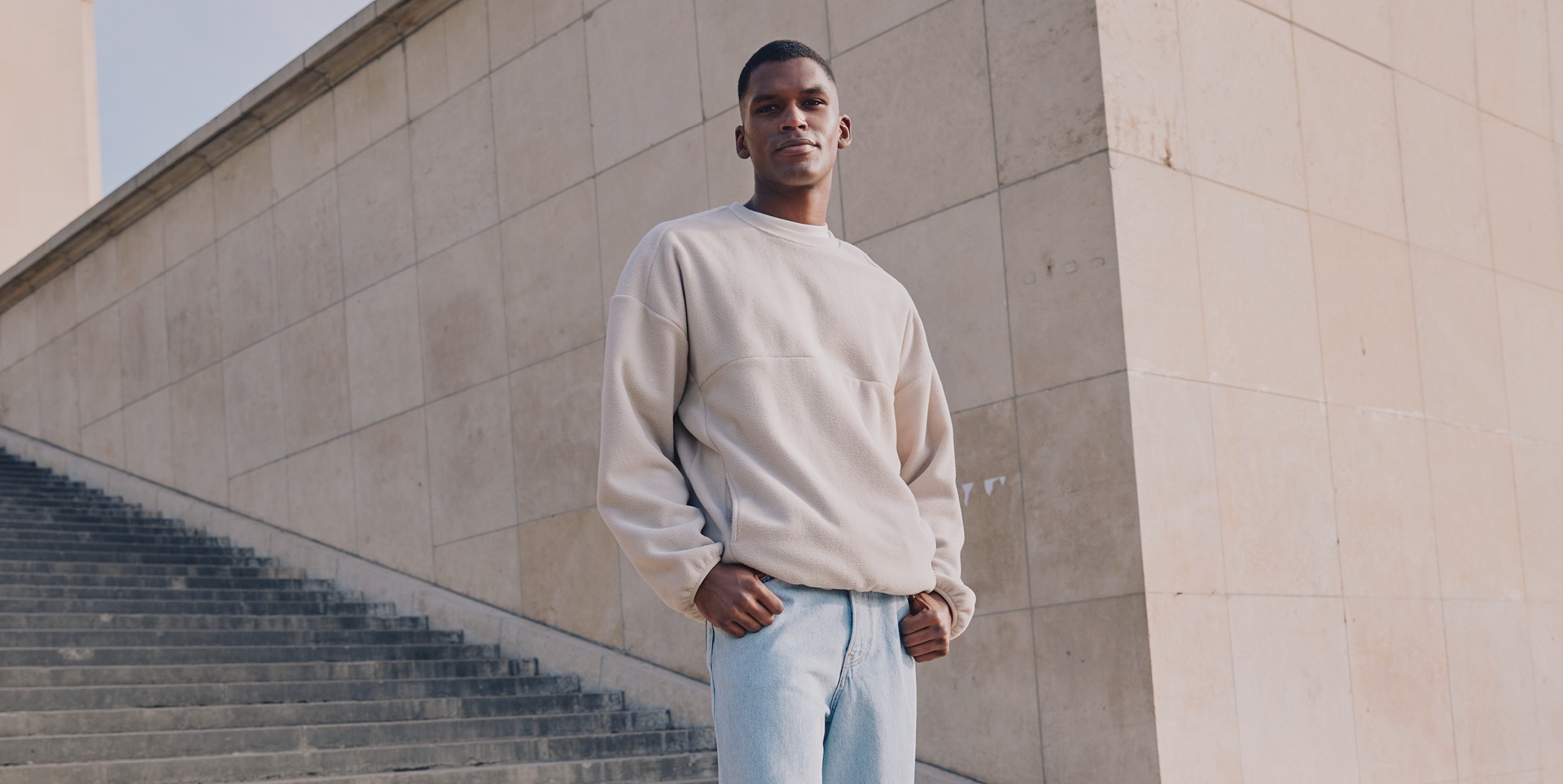 Ein Mann posiert in einem beigen Sweatshirt und blauen Jeans an einer Treppe vor einer Wand. Ein Mann posiert in einem beigen Sweatshirt und blauen Jeans an einer Treppe vor einer Wand.