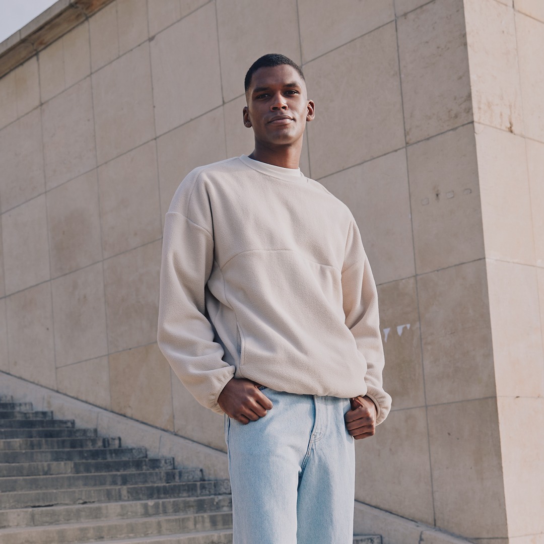 Ein Mann posiert in einem beigen Sweatshirt und blauen Jeans an einer Treppe vor einer Wand. Ein Mann posiert in einem beigen Sweatshirt und blauen Jeans an einer Treppe vor einer Wand.