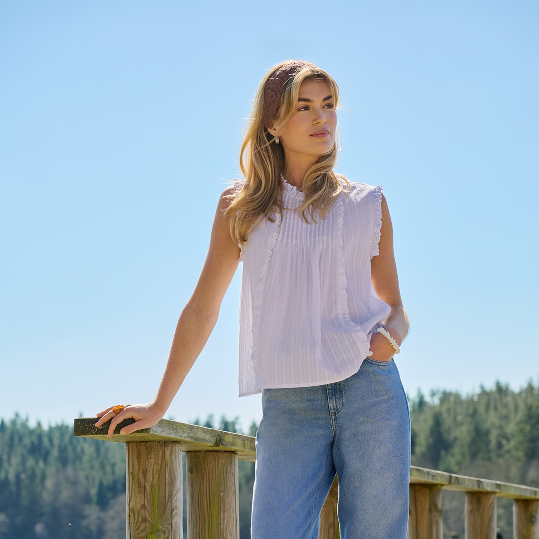 Eine Frau mit langen Haaren trägt ein weißes Oberteil und ein Stirnband, vor einem blauen Himmel.