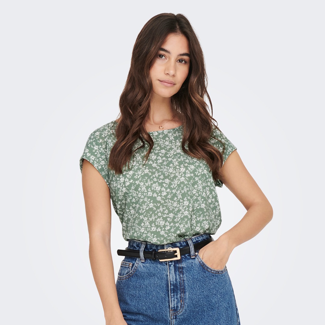 Eine junge Frau trägt ein grün mit Blumenmuster bedrucktes Oberteil und eine blaue Jeans.